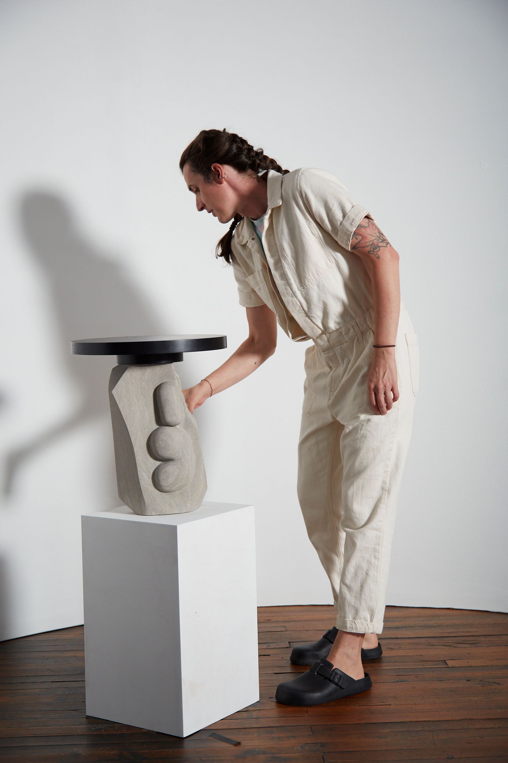 The androgynous artist touches a stone table on a plinth.